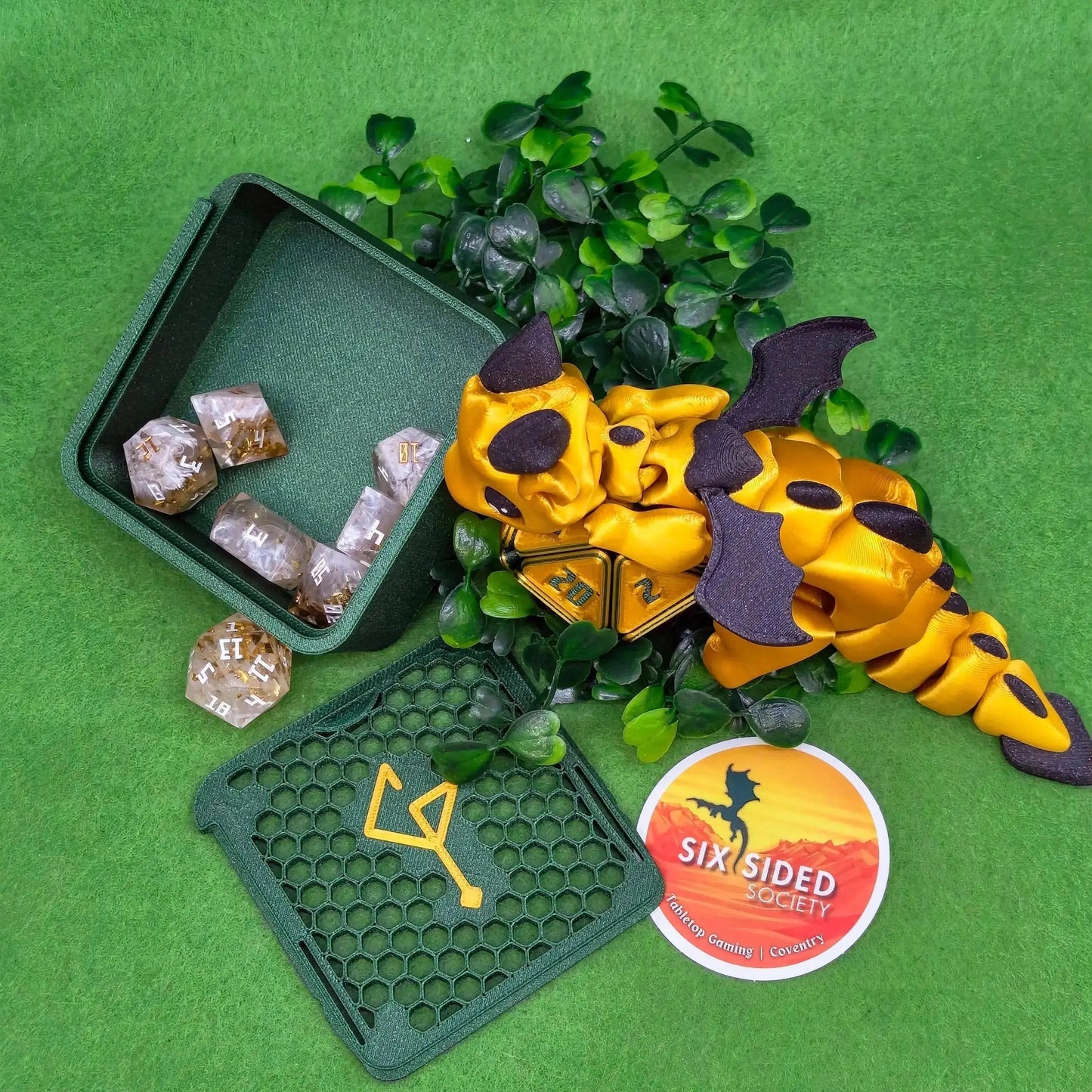 Yellow dragon-shaped dice with purple accents on a green surface with dice and a branded mat. 3D-printed D20 dice trinket box by Six Sided Society, tabletop gaming storage for Dungeons & Dragons, RPG dice organizer, eco-friendly gift box for dice collectors and dungeon masters, perfect accessory for board games and role-playing sessions