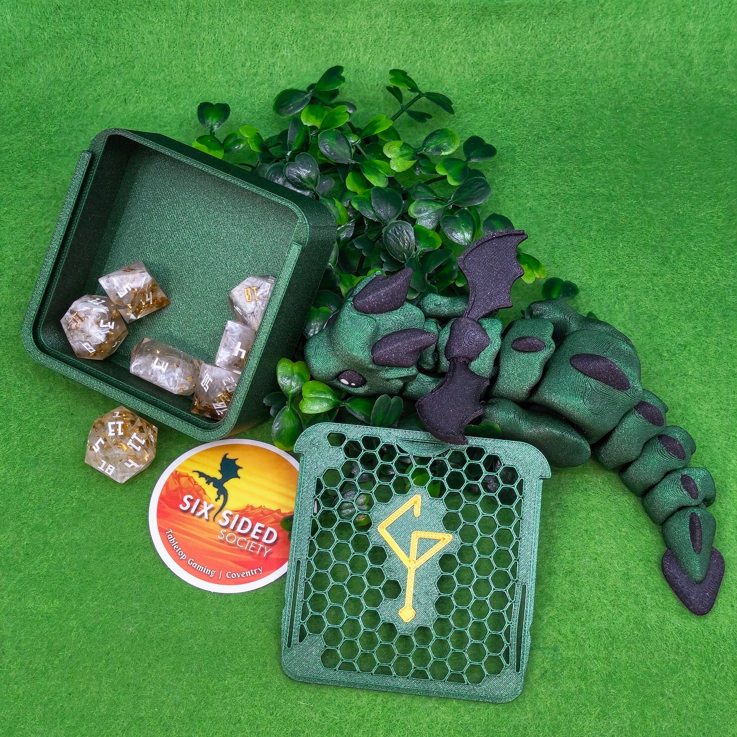 Green dragon toy with dice and a box on a green background. 3D-printed D20 dice trinket box by Six Sided Society, tabletop gaming storage for Dungeons & Dragons, RPG dice organizer, eco-friendly gift box for dice collectors and dungeon masters, perfect accessory for board games and role-playing sessions