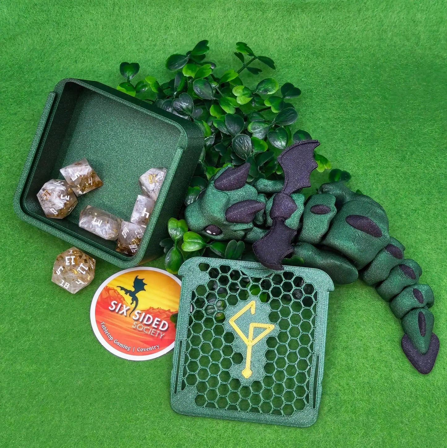 Green dragon toy with dice and a box on a green background. 3D-printed D20 dice trinket box by Six Sided Society, tabletop gaming storage for Dungeons & Dragons, RPG dice organizer, eco-friendly gift box for dice collectors and dungeon masters, perfect accessory for board games and role-playing sessions